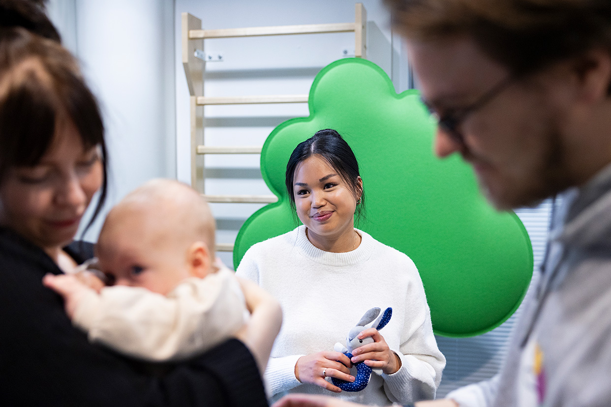 Kaksi henkilöä pitelee vauvaa ja takana seisoo opiskelija