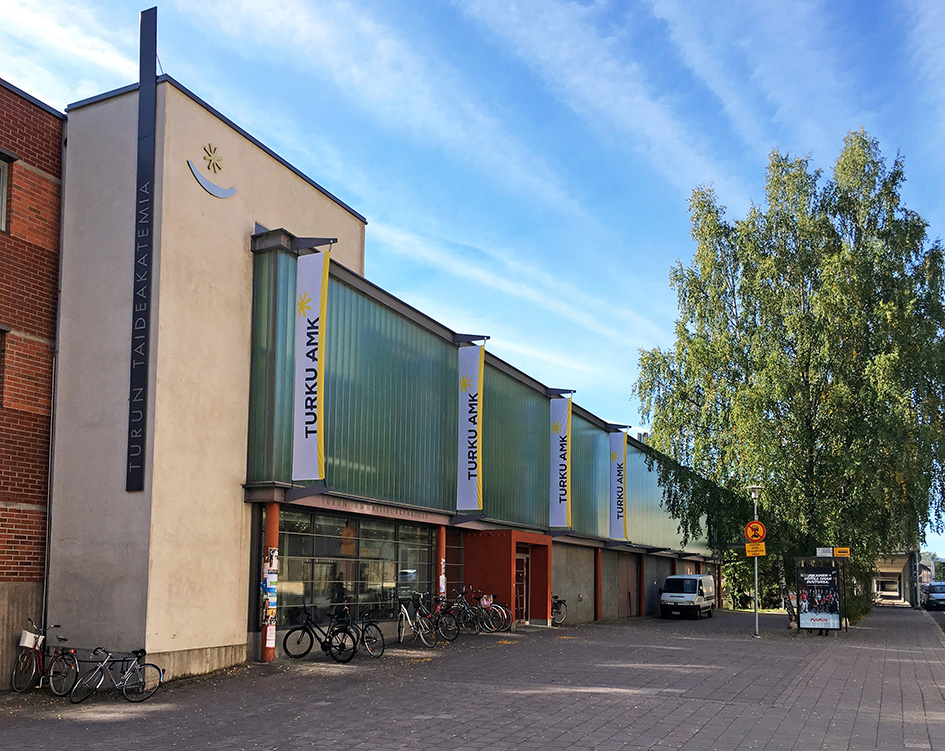 Kampusrakennus kuvattuna kesäaikaan Linnankadun puolelta. Koivu, bussipysäkki ja pysäköityjä autoja kadun varressa.