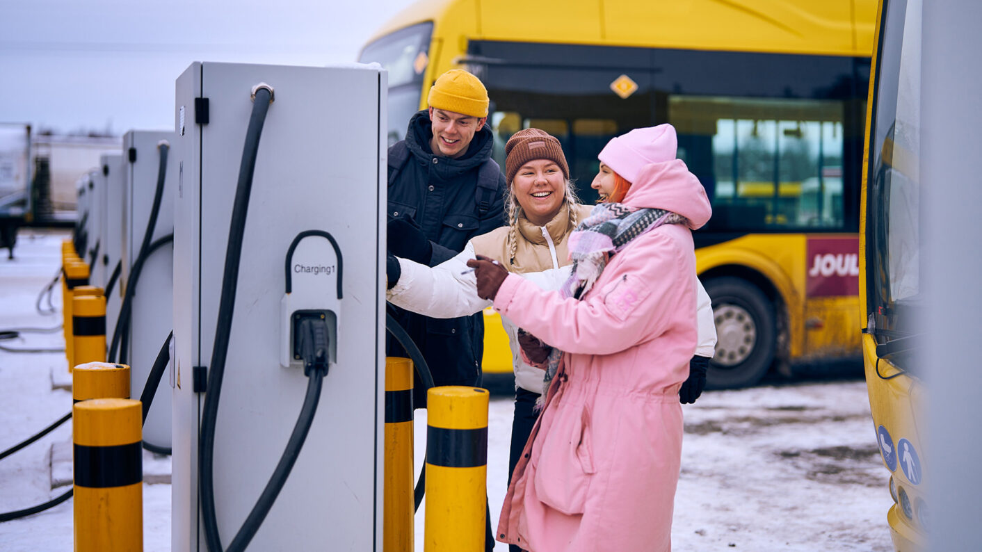 Kolme ihmistä tutkii bussin latauslaitetta hymyillen, taustalla keltainen bussi, maassa on lunta.