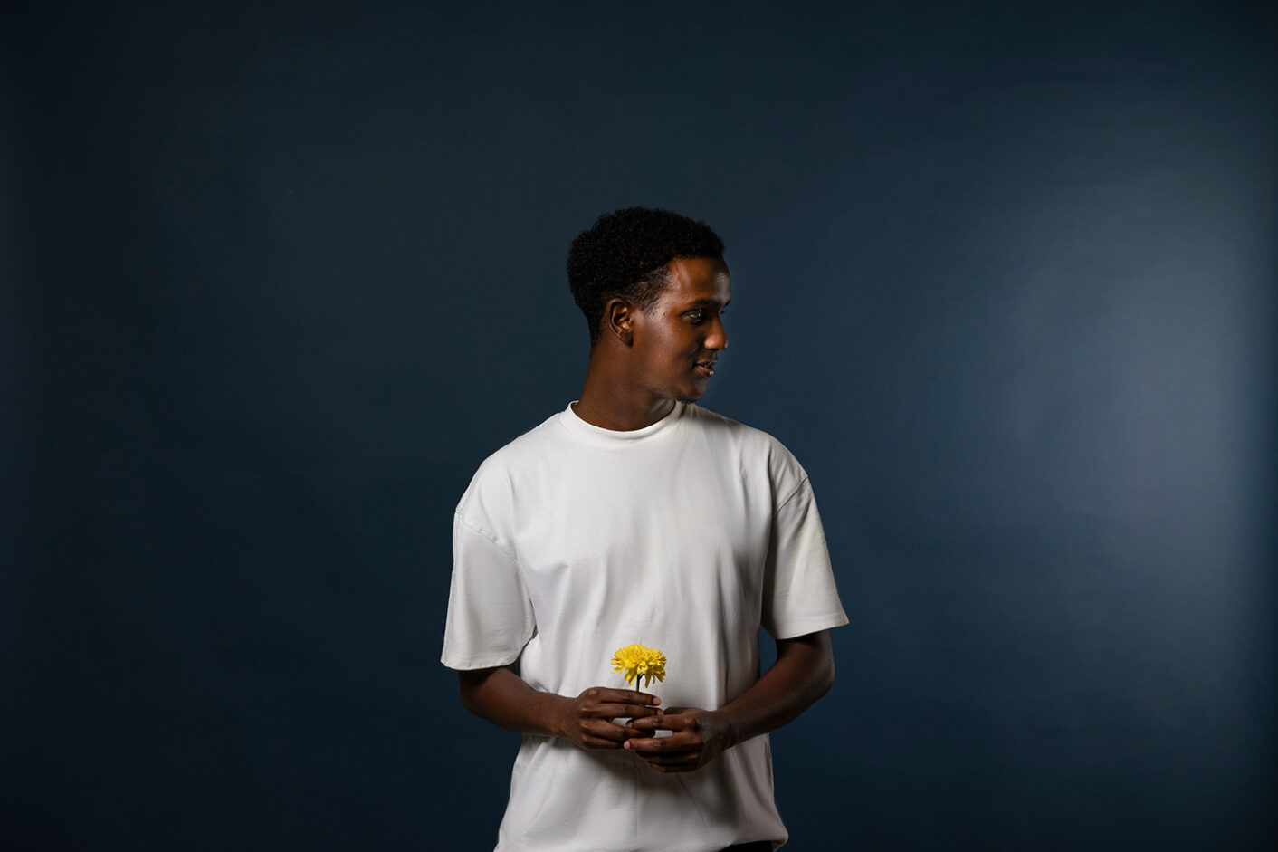 Person is standing in front of a dark blue background. They is holding a yellow flower in theri hands.