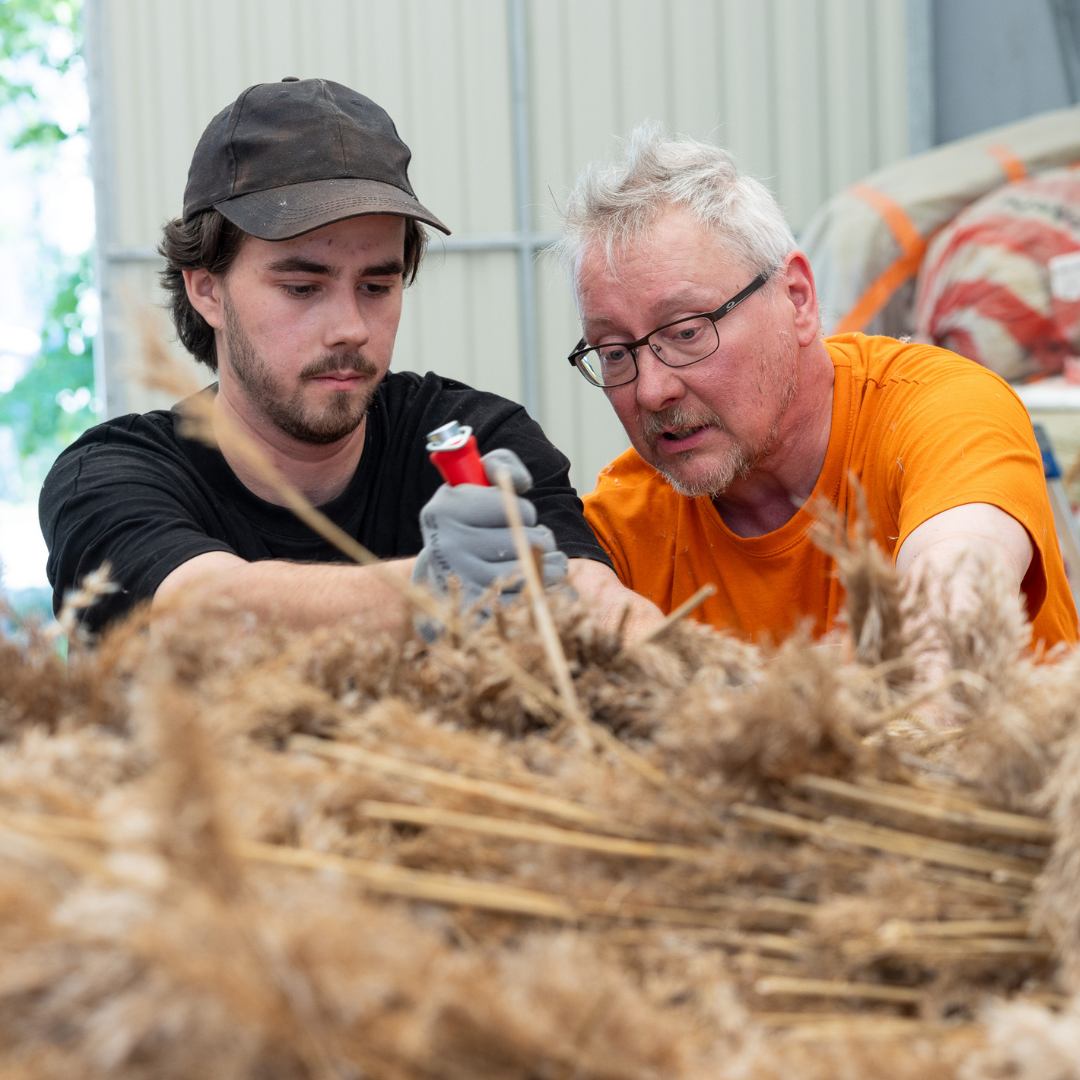 Two people work closely on the reed roof element.